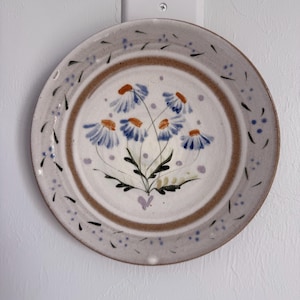 May include: A round ceramic plate with a speckled off-white glaze featuring a hand-painted bouquet of blue and orange daisy-like flowers in the center. The rim is decorated with a brown band, small blue dots, and dark green leaf-like markings.