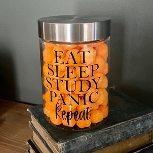 May include: A glass jar with a silver lid filled with orange cheese puffs. The jar has a black vinyl sticker with the text "Eat Sleep Study Panic Repeat".