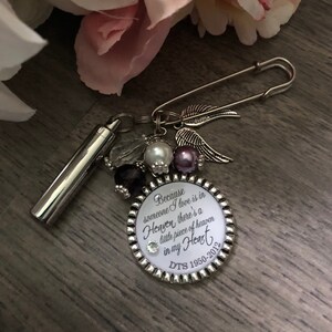 May include: A silver charm bracelet with a round charm that reads "Because someone I love is in Heaven, there's a little piece of heaven in my heart. DTS 1950-2012". The charm is surrounded by small purple and white beads, a silver angel wing charm, and a silver safety pin.