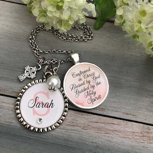 May include: Silver necklace with a charm that says "Confirmed in Christ Blessed by God Guided by the Holy Spirit" and a charm with the letter "S" and the name "Sarah" on it.