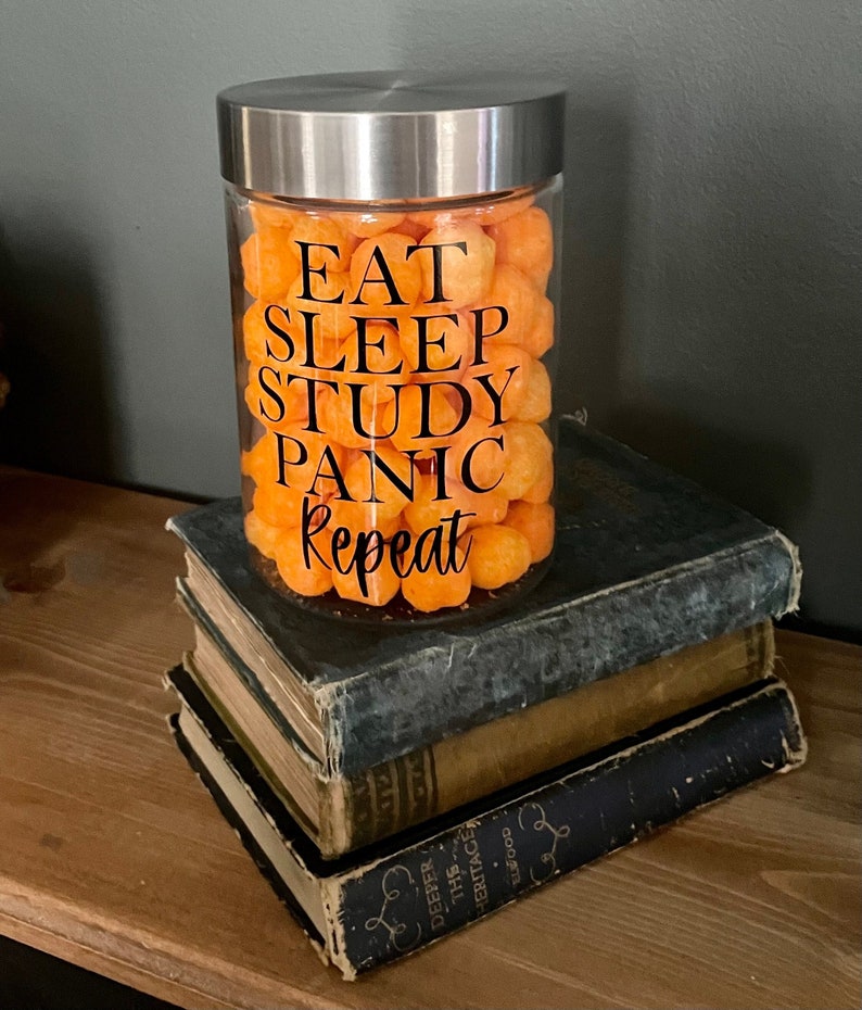 May include: A glass jar with a silver lid filled with orange cheese puffs. The jar has a black vinyl sticker with the text "Eat Sleep Study Panic Repeat".