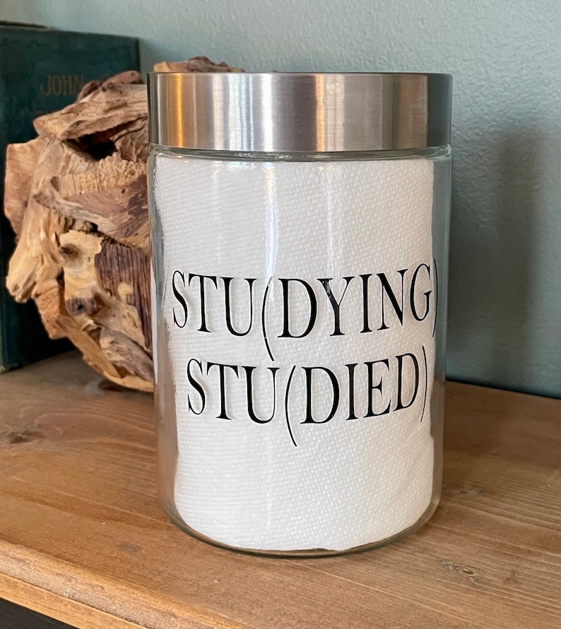 May include: A clear glass jar with a silver lid and a white paper towel inside. The jar has a black vinyl sticker with the text "STU(DYING) STU(DIED)"