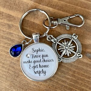 May include: A silver keychain with a compass charm, a blue crystal charm, and a round charm with the text "Sophie, Have fun make good choices & get home safely".