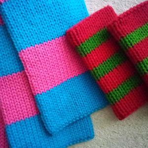 May include: A hand-knitted scarf featuring wide horizontal stripes in bright pink and cyan blue, accompanied by two matching hand-knitted wrist warmers or cuffs that display alternating horizontal stripes in vibrant red and grass green colors.
