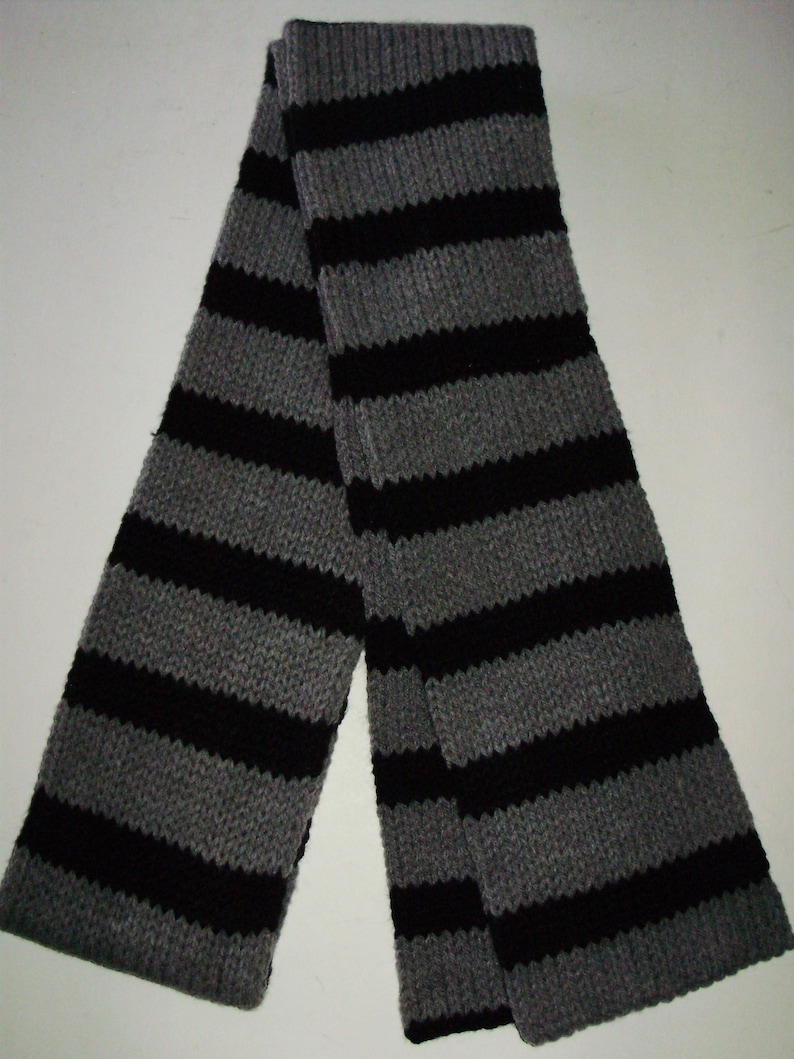 May include: A black and gray striped knitted scarf. The scarf is folded in half and is lying flat on a white surface.
