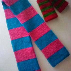 May include: A long knitted scarf featuring horizontal stripes in bright pink and cyan blue, accompanied by two smaller rectangular knitted wrist warmers or cuffs that display horizontal stripes in alternating red and green colors.