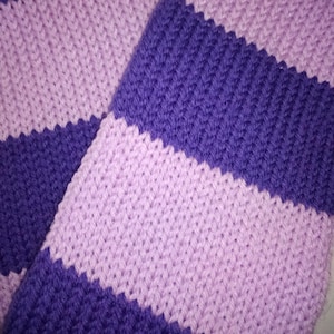May include: Close-up of a knitted scarf with alternating stripes of light purple and deep purple. The scarf is made of a soft, textured yarn, and the stripes run horizontally across the fabric. The knitted pattern is visible.