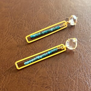 May include: A pair of gold earrings with a rectangular frame and a row of blue gemstones. Each earring has a clear crystal at the bottom.