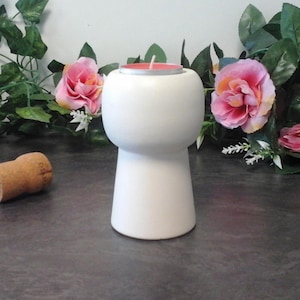 Peut inclure: Un bougeoir en céramique blanc mat en forme de base de bouchon de champagne avec un sommet arrondi et une base plate. Une petite bougie ronde avec une surface en cire rose et une mèche blanche est insérée dans l'ouverture supérieure du support.
