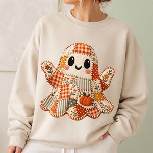 Op de afbeelding: Een beige sweater met ronde hals en een grote, centrale afbeelding van een geest gemaakt van verschillende patchworkpatronen. Het ontwerp van de geest bevat oranje en witte ruitjes, bloemenprints, stippen en een kleine oranje pompoen.