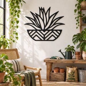 May include: A black metal wall art piece featuring a stylized succulent plant in a geometric patterned pot. The design consists of sharp, pointed leaves extending upward from a semi-circular base decorated with a repeating chevron pattern.