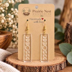 Pode incluir: Um par de brincos retangulares de acrílico transparente com um design de fases da lua impresso em branco. Os brincos pendem de ganchos de metal dourado e estão presos a um cartão de papelão bege com o texto Prairie Nest Woodworking.