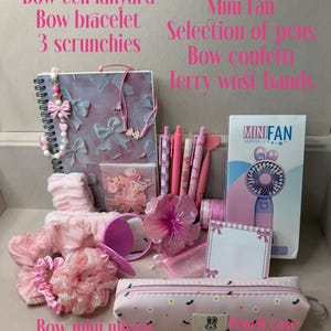 May include: A collection of pink stationery and accessories including a spiral notebook, cell lanyard, bracelet, three scrunchies, pens, a mini fan in packaging, a hibiscus hair clip, a mirror, a pencil case, post-its, and washi tape.