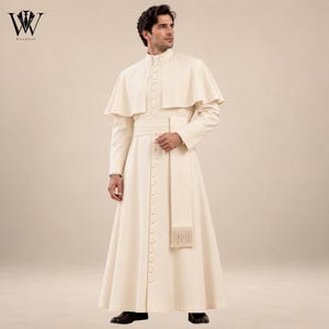 May include: A floor-length cream-colored cassock featuring a short capelet, a high collar, and a central row of buttons extending from the neck to the hem. The garment includes a wide matching sash with a fringed end and long, fitted sleeves.