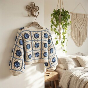 May include: A long-sleeved crochet jumper constructed from cream-coloured square panels, each featuring a central blue floral motif. The garment has a ribbed hem and cuffs, with a square neckline, showcasing a detailed handmade yarn texture.