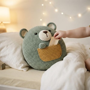 Può includere: Un cuscino rotondo all'uncinetto a forma di orso color verde salvia, con muso color crema, naso ricamato marrone scuro e due piccole orecchie. L'orso presenta una tasca all'uncinetto color senape sul davanti, contenente un cartoncino beige.