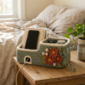 May include: A sage green crocheted storage caddy with a dedicated slot for a smartphone, a charging cable port, and a side compartment. The exterior is decorated with three-dimensional crocheted flowers in rust and pink with green leaves.