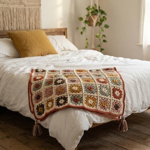 Puede incluir: Una manta de ganchillo hecha a mano con una cuadrícula de motivos cuadrados en tonos apagados de amarillo mostaza, verde salvia, rosa empolvado y crema. La manta está rematada con un borde festoneado y borlas decorativas en las esquinas.