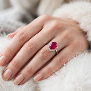 May include: A silver ring featuring a large, oval-cut, deep red ruby gemstone as the centrepiece. The ruby is surrounded by a halo of small, clear, round-cut white diamonds set into a polished silver band, creating a classic and elegant appearance.