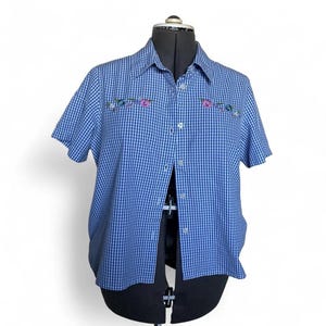 Op de afbeelding: Een overhemd met korte mouwen en knoopsluiting, voorzien van een blauw-wit vichy-ruitpatroon. Het overhemd heeft een klassieke kraag en een knoopsluiting aan de voorzijde. Geborduurde bloemdetails in roze, groen en wit sieren de borst.