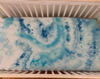 May include: A white crib with a blue and white tie-dye fitted sheet. The sheet has a swirling pattern of light and dark blue.