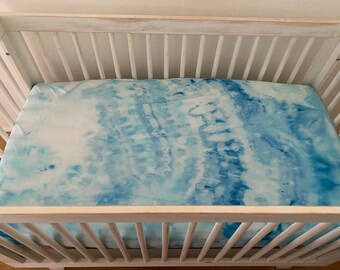 May include: A white wooden crib with a blue and white tie-dye fitted sheet. The sheet has a swirling pattern of light and dark blue.
