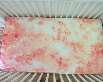 May include: A white crib sheet with a tie-dye pattern in shades of pink and orange.