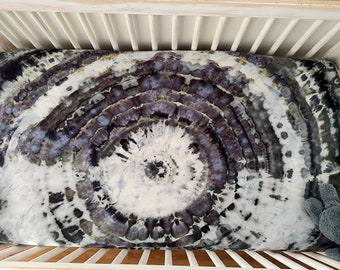 May include: A white, gray, and black tie-dye fitted crib sheet with a circular pattern. The sheet is in a white crib with wooden slats.