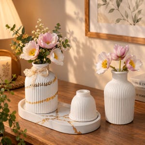 May include: Two white ribbed ceramic vases and a small ribbed bowl on an oval white tray with gold veining. One vase holds pink and white flowers with a beige ribbon tied around the neck. The other vase holds three pink flowers.