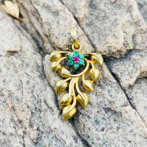 May include: A gold pendant featuring a cluster of small green stones arranged in a flower shape with a single pink stone at the center. The pendant is designed with multiple gold leaf-shaped elements extending downward from the flower.