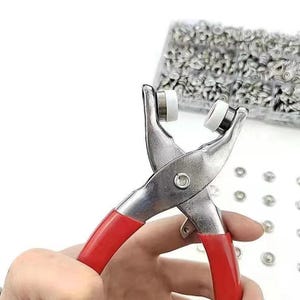 May include: A metal snap fastener plier tool with bright red rubberized handles and white circular press heads, positioned near a clear plastic storage container filled with numerous small silver-colored metal snap buttons.