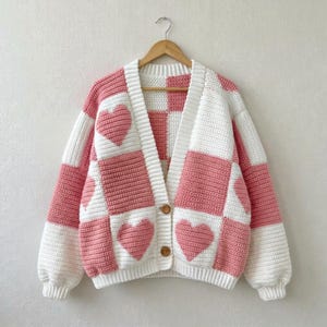 May include: A long-sleeve crochet cardigan featuring a pink and white checkered pattern with integrated heart motifs. The garment has a V-neckline, a button-front closure with two brown buttons, and ribbed cuffs, all displayed on a wooden hanger.
