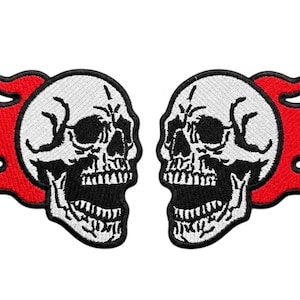 Puede incluir: Dos parches bordados que presentan calaveras blancas con detalles en negro y bocas abiertas, cada una rodeada por llamas de color rojo vibrante con contornos negros. Las calaveras están orientadas frente a frente, creando un diseño simétrico.