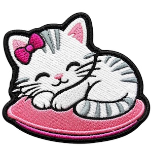 Puede incluir: Un parche bordado que muestra un gatito blanco con rayas grises durmiendo sobre un cojín rosa. El gatito tiene un lazo rosa brillante en la cabeza, los ojos cerrados y una pequeña nariz rosa. El diseño está delineado con un borde negro grueso.