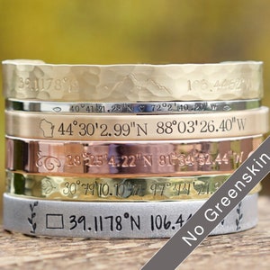 May include: A stack of six metal cuff bracelets in various finishes, including gold, silver, and copper. Each bracelet is engraved with coordinates and decorative elements. The bracelets are stacked on top of each other.