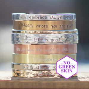 May include: A stack of metal bracelets in various finishes, including silver, gold, and copper. Each bracelet is engraved with an inspirational phrase, such as "embrace change" and "Life is good". The bracelets are stacked on top of each other.