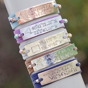 May include: A stack of personalized bracelets with suede bands in various colors. Each bracelet features a metal plate engraved with latitude and longitude coordinates and a state outline. The bracelets are stacked on a light gray surface.