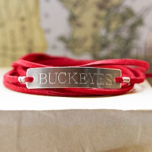 May include: A red leather wrap bracelet with a silver rectangular tag engraved with the word "BUCKEYES".