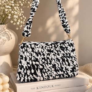 May include: A rectangular handbag crafted from thick, black and white crocheted yarn. The bag features a matching braided shoulder strap attached with gold-toned metal clasps. The crochet pattern creates a dense, textured surface throughout the body.