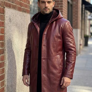 May include: A long, deep burgundy leather coat featuring a hood with a dark interior lining, a button-front closure, and two side pockets. The coat is worn over a black turtleneck jumper, providing a structured look with visible seam detailing.