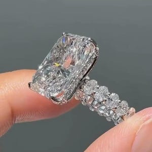 Op de afbeelding: Een verlovingsring in radiant-slijpvorm met een grote rechthoekige middensteen met een briljant gefacetteerd oppervlak. De ringband is gemaakt van gepolijst zilverkleurig metaal en is bezet met meerdere kleine ronde stenen langs de zichtbare schacht.