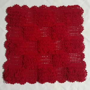 Puede incluir: Un tapete cuadrado de ganchillo rojo con un patrón alterno de bloques texturizados sólidos y secciones de rejilla abierta. Los bordes están rematados con un borde festoneado, creando una pieza textil decorativa de color rojo intenso.
