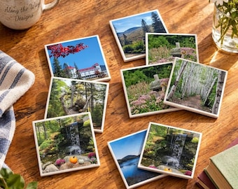 May include: A set of square coasters featuring various nature and landscape photographs, including a waterfall, a forest path, a mountain range, and a building, arranged on a wooden surface with a mug and a glass vase.
