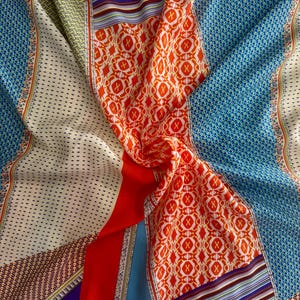 May include: A multi-colored scarf featuring a patchwork design with vibrant orange, blue, red, and cream sections. The fabric displays various geometric patterns, including small dots, intricate cross motifs, and horizontal stripes.
