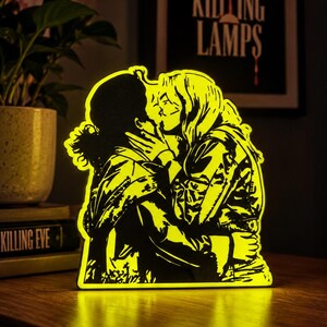 May include: A bright yellow LED light display featuring a silhouette of two people kissing. The design shows one person with curly hair and another with long hair, both wearing jackets. The light emits a vibrant glow, highlighting the detailed outlines of the figures against the dark background.