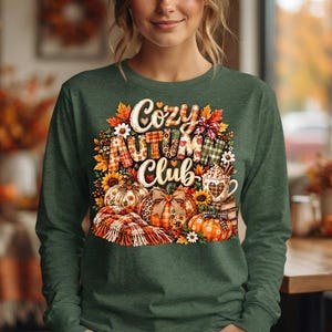 May include: A long-sleeve olive green shirt featuring a graphic print with the text Cozy Autumn Club. The design includes orange pumpkins, sunflowers, a plaid blanket, and a mug of hot cocoa surrounded by colorful autumn leaves and patterns.