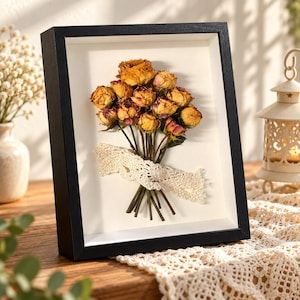 Può includere: Una cornice a cassetta rettangolare nera contenente un bouquet pressato di rose essiccate gialle e rosa. Gli steli sono legati insieme con un nastro di pizzo all'uncinetto color crema su uno sfondo bianco all'interno della cornice.