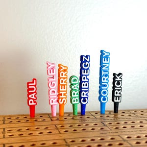Peut inclure: Un ensemble de sept pions de cribbage en plastique coloré insérés dans un plateau de jeu en bois. Les pions sont rouge, rose, orange, vert, bleu, bleu clair et noir, chacun portant une étiquette verticale : Paul, Ridgley, Sherry, Brad, Cribpegz, Courtney et Erick.