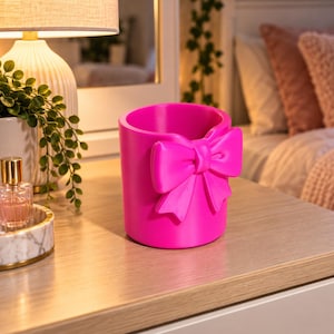 May include: A vibrant hot pink cylindrical container featuring a prominent, three-dimensional bow attached to the front exterior. The object has a smooth, solid-coloured finish and an open top, designed as a decorative vessel for small items.