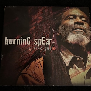May include: A square music album cover featuring a portrait of a man with dreadlocks and a beard. The text reads burning spear, LIVING DUB, and Volume 4. The background shows a dark brick wall with red lighting accents, creating a high-contrast visual.
